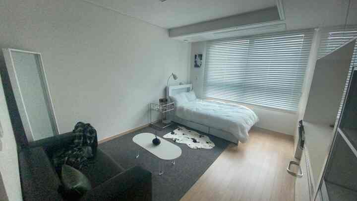 White and gray toned bedroom featuring a single bed, blinds covering the window, a gray sofa, and a spotted rug, creating a simple and calm atmosphere