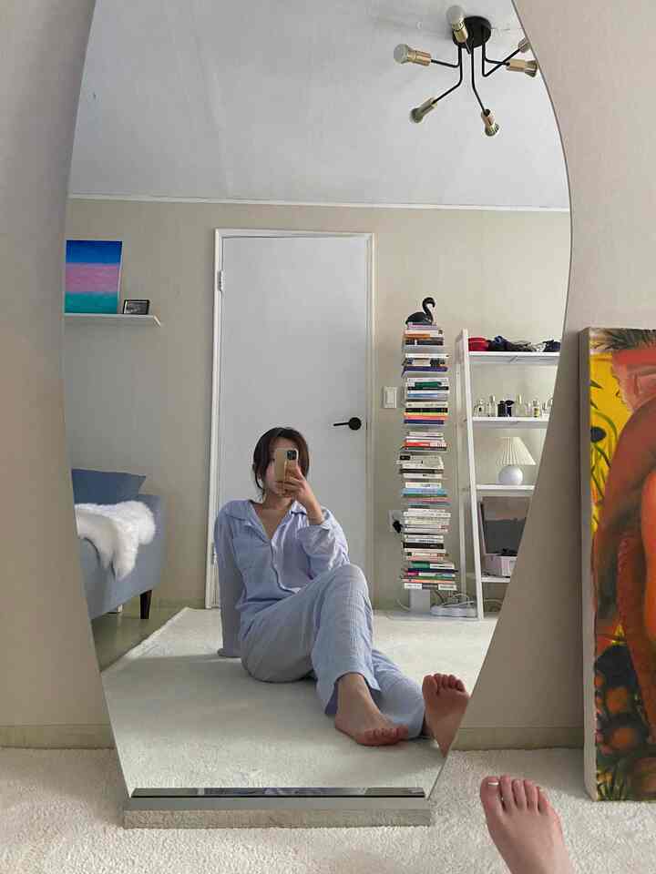 A white and beige toned living room featuring a large wall mirror, bookshelf, and sofa with a simple and cozy atmosphere