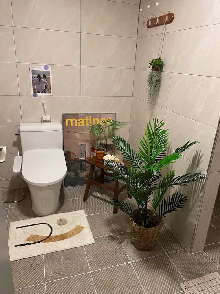 ベージュの壁と床タイルのナチュラルトーンのトイレ、中央に白い便器があり、木目調のスツールと植物が配置された雰囲気のあるインテリア