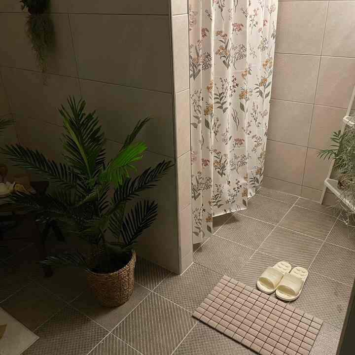 Beige-toned bathroom featuring a large green plant, floral shower curtain, diatomaceous earth bath mat, and white bathroom slippers creating a natural ambiance