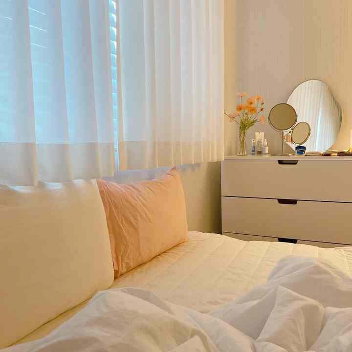 A white and beige toned bedroom featuring curtains with blinds, pillows, bedding, a dresser, and a vase with flowers, creating a cozy atmosphere.