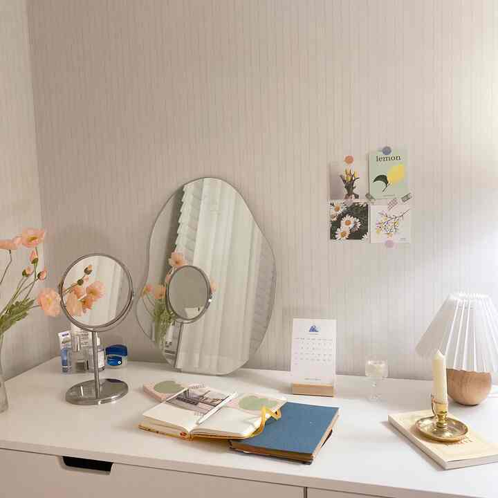 Soft beige and white toned room featuring dresser with table mirrors, vase with flowers, and candle, creating a natural vanity space