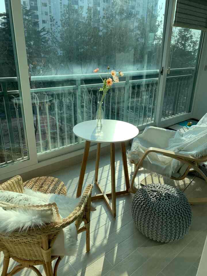 自然光が差し込むベージュと白のトーンのバルコニー空間、ラタンの椅子と丸テーブルが配置された雰囲気のあるカフェ風インテリア