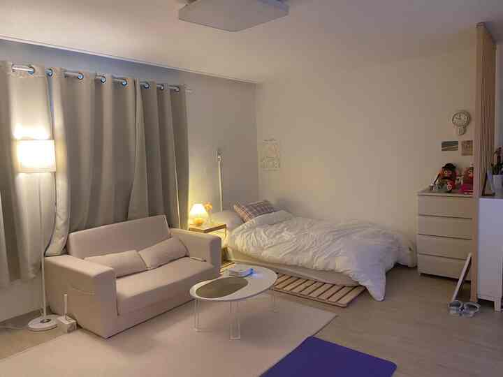 ホワイトとベージュのトーンのワンルームリビング、ベッドとソファ、カーテンが配置されたシンプルで清潔感のあるインテリア