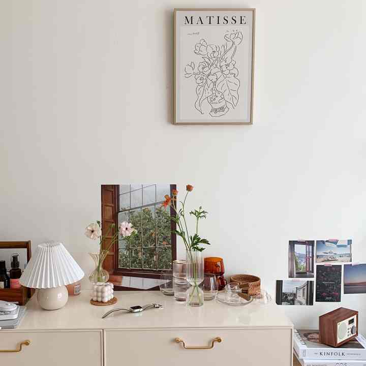 A bright white-walled room featuring a natural brown dresser, poster, vases, and a table lamp creating a simple and cozy interior