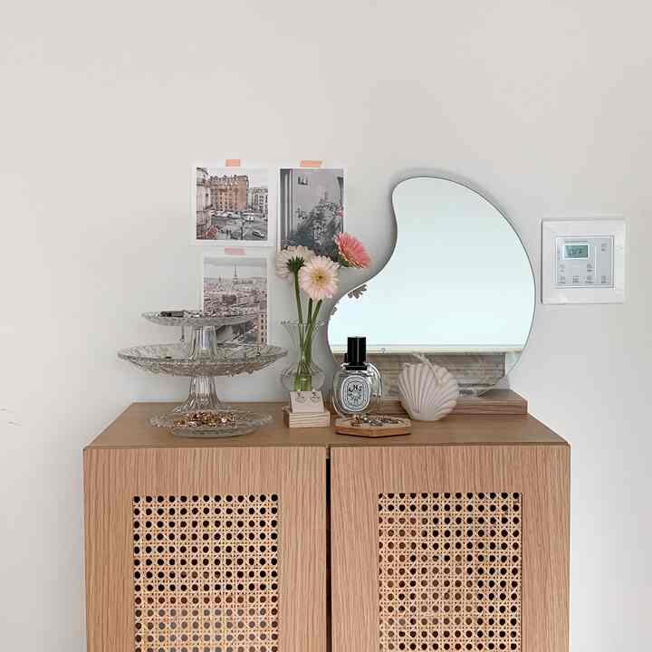 A cozy space with white walls and wood cabinet, featuring an irregular shaped mirror, vase with flowers, and accessories on a natural vanity setup