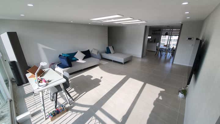 A white and light gray toned living and dining space featuring a sofa, desk, and large TV with a simple modern interior