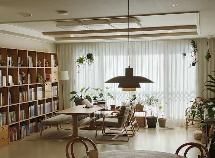 明るいナチュラルモダン調のリビング・ダイニングスペース、木製の本棚とダイニングテーブル、カーテン、多様な植物が配置された温かみのあるインテリア