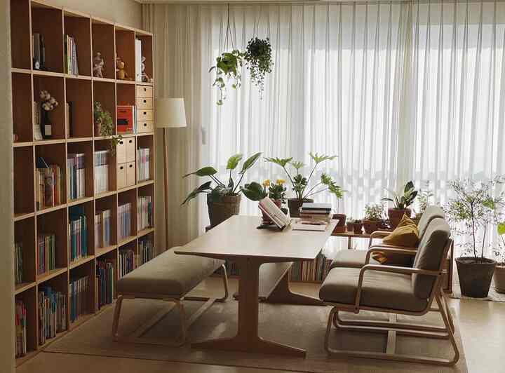 ナチュラルな木目調と白いカーテンのリビングには、本棚や植物、くつろげる椅子が配置された落ち着いた読書空間