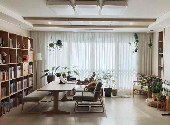 Natural-toned living room study featuring a large bookshelf, wooden dining table, and abundant plants creating a cozy interior