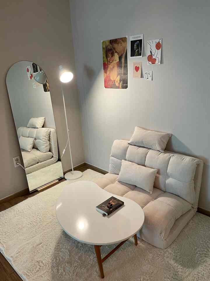 ベージュとクリーム色調のコンパクトなリビング、ソファベッドとフロアランプ、ラグ、鏡、ポスターが配置されたシンプルなインテリア