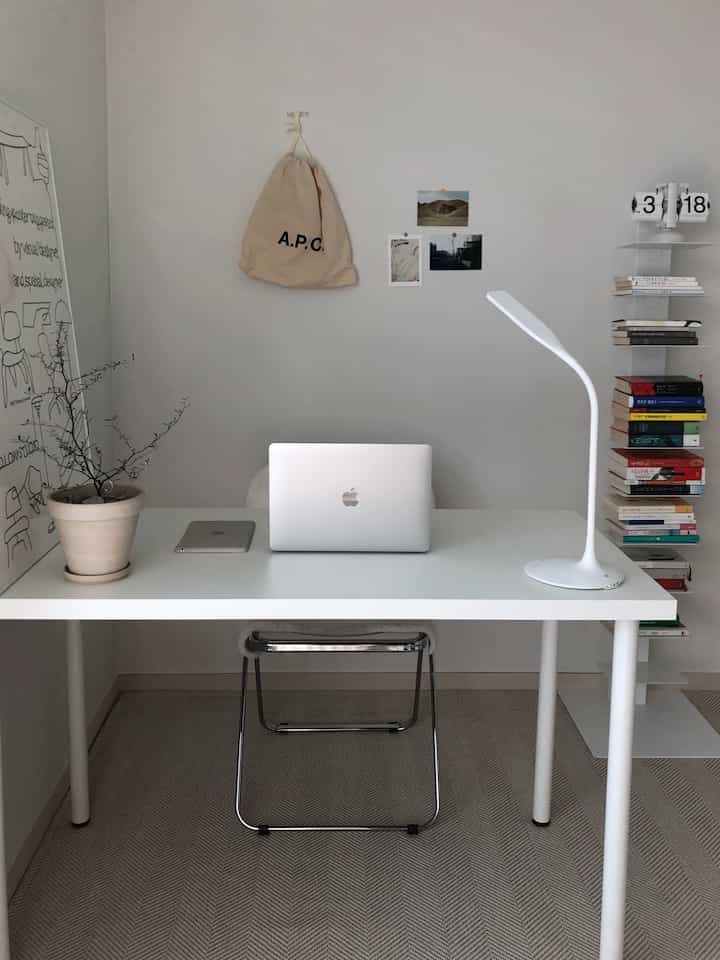 Simple white-toned study space featuring a white desk with laptop and lamp centered, and a bookshelf on the wall for a minimal interior