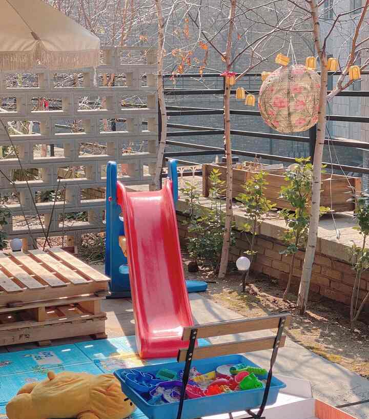 パステル調の屋外テラス空間、子供用滑り台とおもちゃ、木製パレットが配置された、赤ちゃんと犬のための温かみのある雰囲気のインテリア