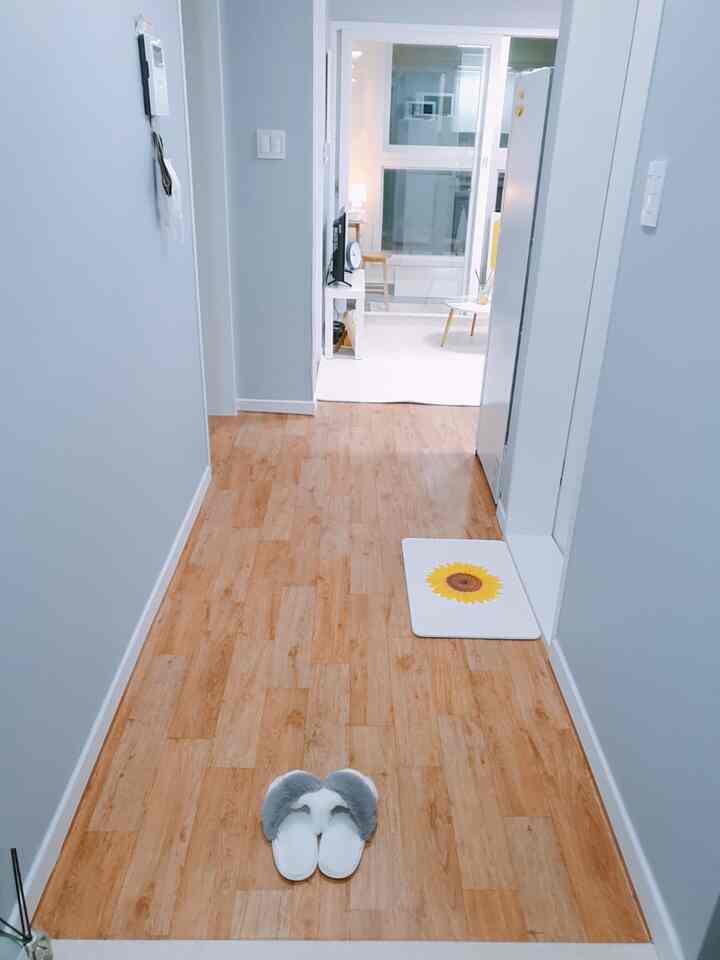 Clean hallway with wood-tone flooring and white walls, featuring gray slippers centrally and a sunflower bath mat near the door on the right