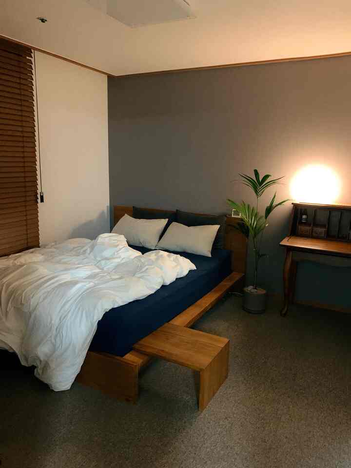 Natural tone bedroom featuring a wooden bed, brown blinds, nightstand, and a green plant, creating a cozy atmosphere