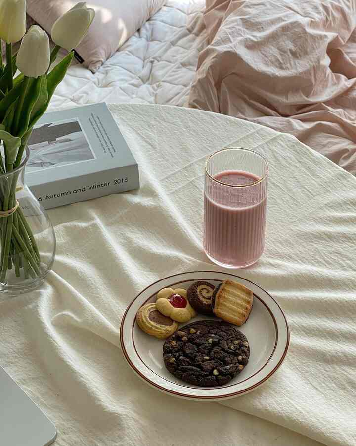 柔らかなベージュとホワイトのトーンのベッドとテーブルクロスの上にクッキーとピンクの飲み物が配置された雰囲気のあるおうちカフェの空間