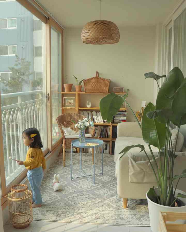 Natural-tone balcony living room featuring a cream sofa, rattan chair, child by the glass door, and lush plants creating a cozy atmosphere