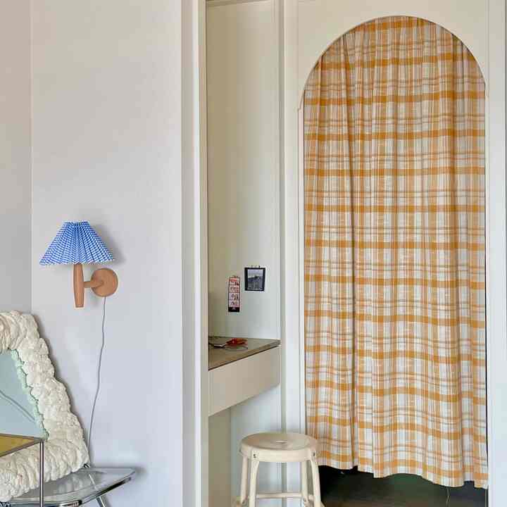 ナチュラルな色調の寝室、黄色のチェックカーテンがかかったアーチ型の入口と壁に青いプリーツ壁照明が配置されたウォークインクローゼットの空間