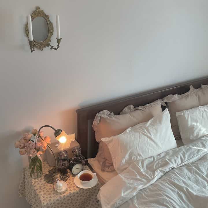 A cozy vintage bedroom featuring white bedding and a brown wooden bed frame, with a vase and table lamp arranged on the nightstand