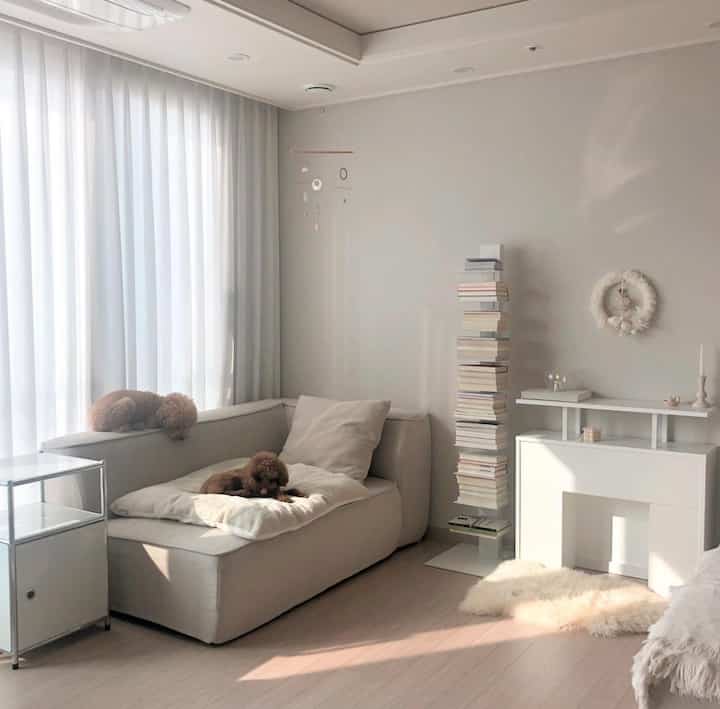 白とベージュの色調のリビング、ベージュのファブリックソファと背の高いブックタワーが配置された温かみのあるシンプルな空間