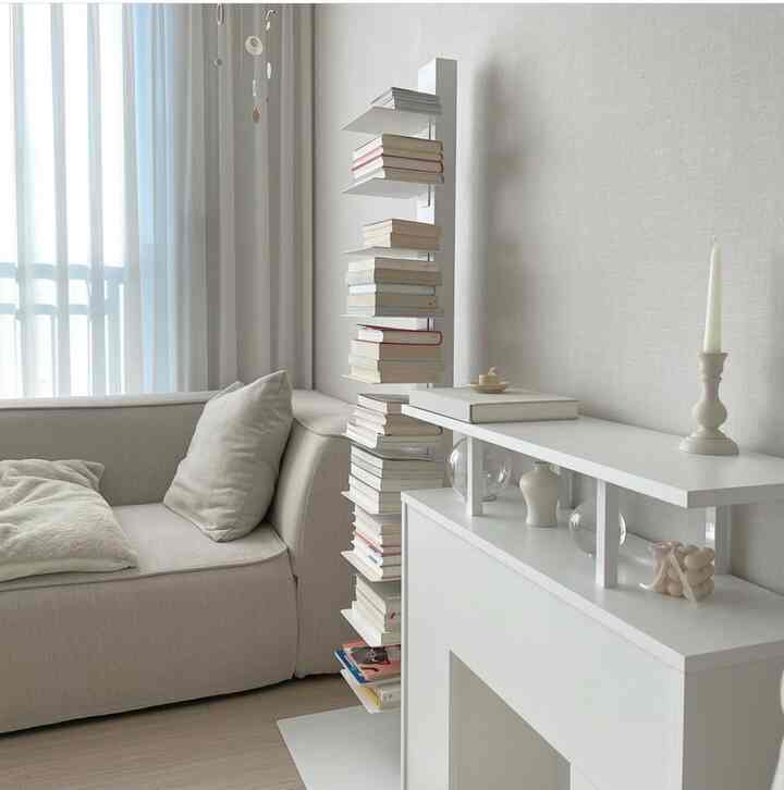 White-toned living room featuring beige sofa, white bookshelf filled with books, fireplace-style console, and hanging mobile in a neat calm space