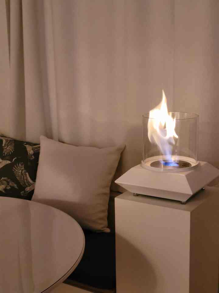 柔らかな白のトーンのワンルーム、遮光カーテンとクッションが配置された落ち着いたエタノール暖炉のある空間