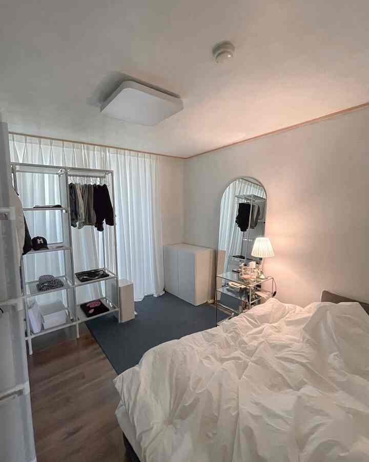 A white and gray toned bedroom featuring large white curtains, storage cabinet, arched full-length mirror, and soft lighting creating a cozy atmosphere