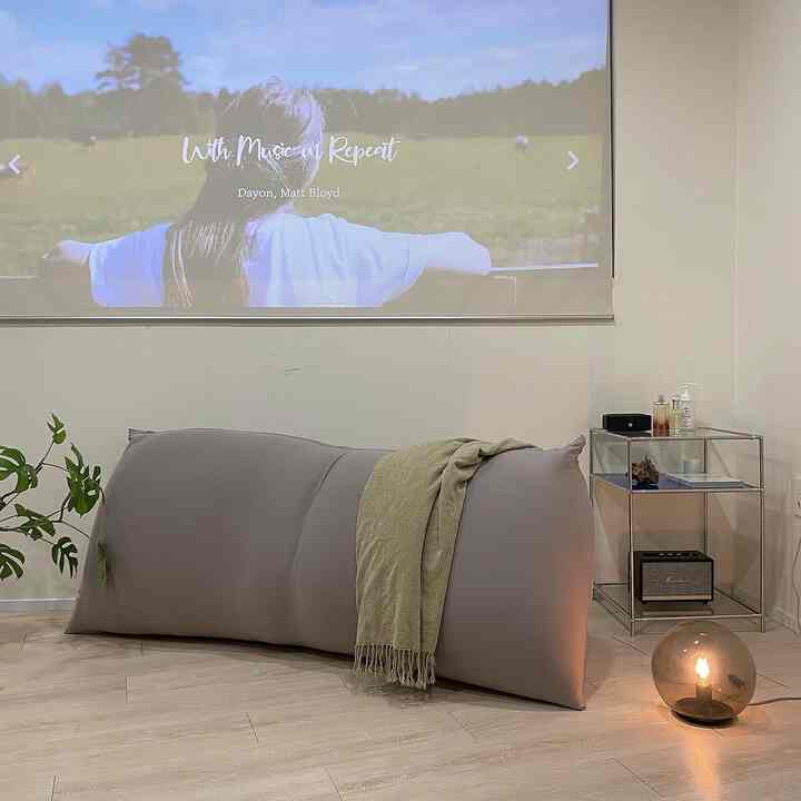 A modern living room in white and beige tones featuring a central Yogibo bean bag with blanket, metal side table and floor lamp to the right
