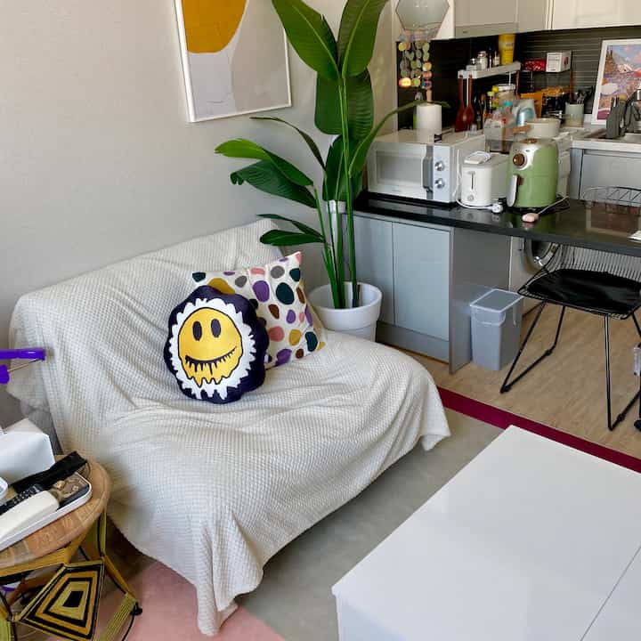 White and natural tone living room featuring sofa with cushions and throw, plant, and adjoining kitchen creating cozy atmosphere