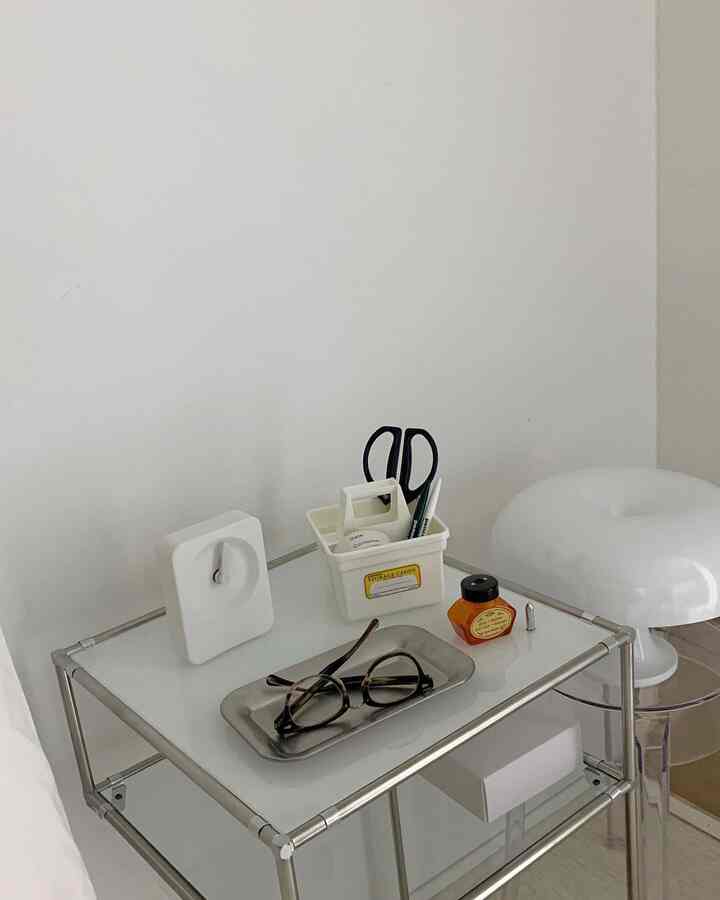 ホワイトとシルバーのトーンの小さな部屋のナイトテーブル、ステンレストレイと時計、文具が整然と配置されたミニマルな空間