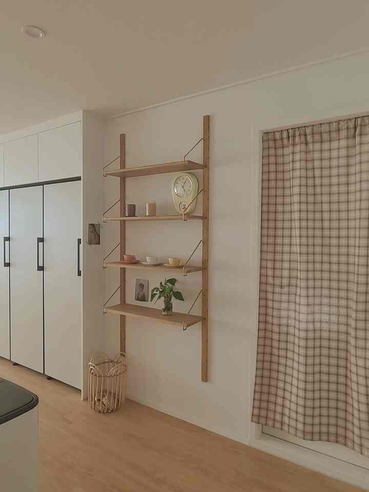 ベージュとホワイトのトーンが特徴のキッチン、チェックカーテンと木製の壁棚、ヴィンテージの時計とティーカップが配置された自然な雰囲気のインテリア