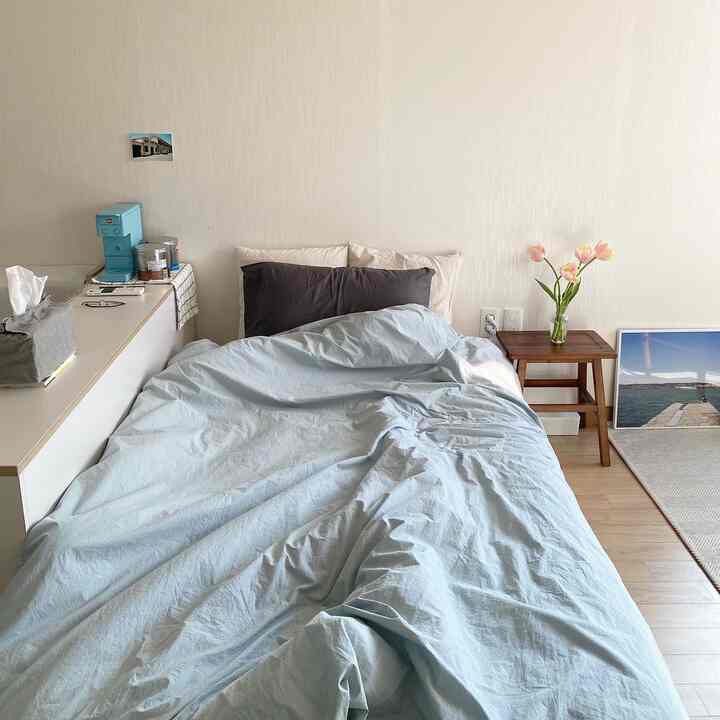 White-walled and wood-toned floor studio bedroom featuring a low bed with pastel blue duvet cover and wooden stool, creating a cozy atmosphere