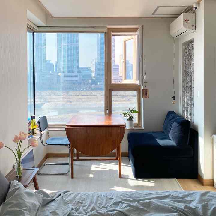 Compact studio apartment with white walls, wood-tone dining table, and navy sofa illuminated by natural light
