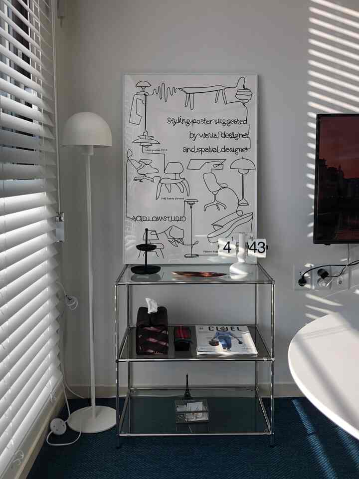 白とネイビーのリビングスペース、左側にウッドブラインドとフロアランプ、中央にガラス製3段シェルフとポスター、小さなフリップクロックが配置されたミニマルなインテリア