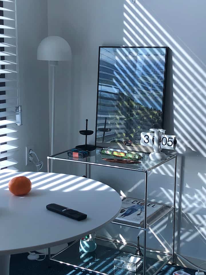 White-toned living room featuring a round table, transparent modular shelf, and wood blinds softly filtering sunlight