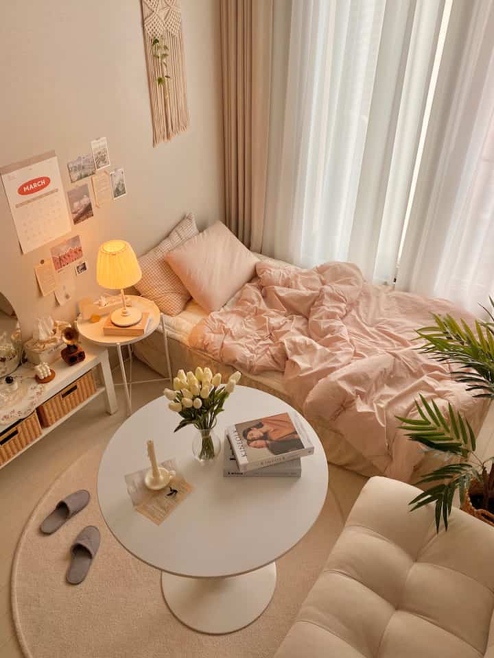 Beige and white toned studio apartment bedroom featuring a cozy bed, round table, and plants in a warm atmosphere