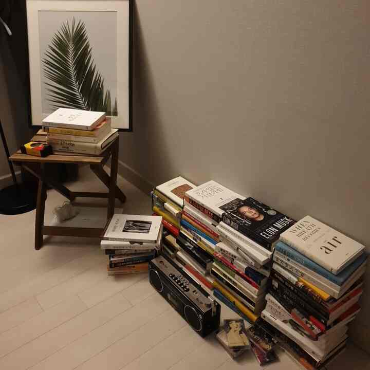 Natural tone bedroom featuring beige walls and floor with stacks of books and a cassette player on the floor, alongside a small wood-toned stool and framed artwork creating a tidy atmosphere
