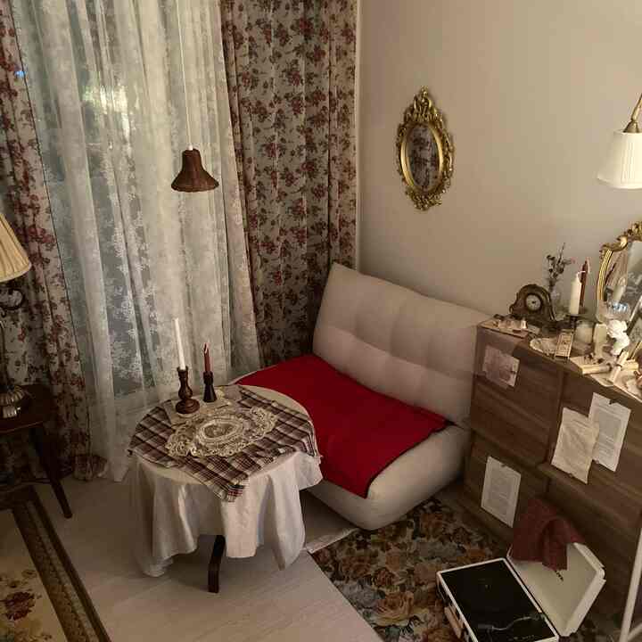 ヴィンテージ調の小さな部屋、ベージュの一人掛けソファと円形テーブル、花柄のカーテンと金色の装飾鏡が配置された雰囲気のあるインテリア