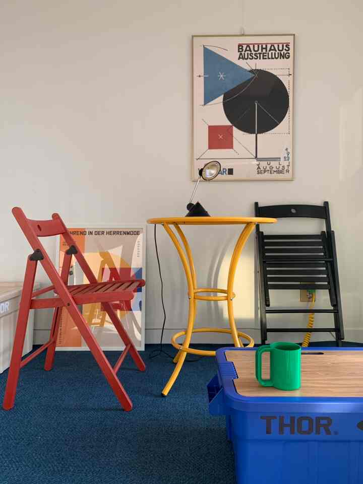 A modern home cafe space featuring a red folding chair, yellow round table, blue rug, and wall posters creating a colorful atmosphere
