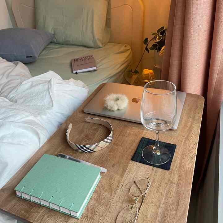 A cozy bedroom with natural wood side table and mint-colored duvet cover, featuring a wine glass and pink curtains beside the bed