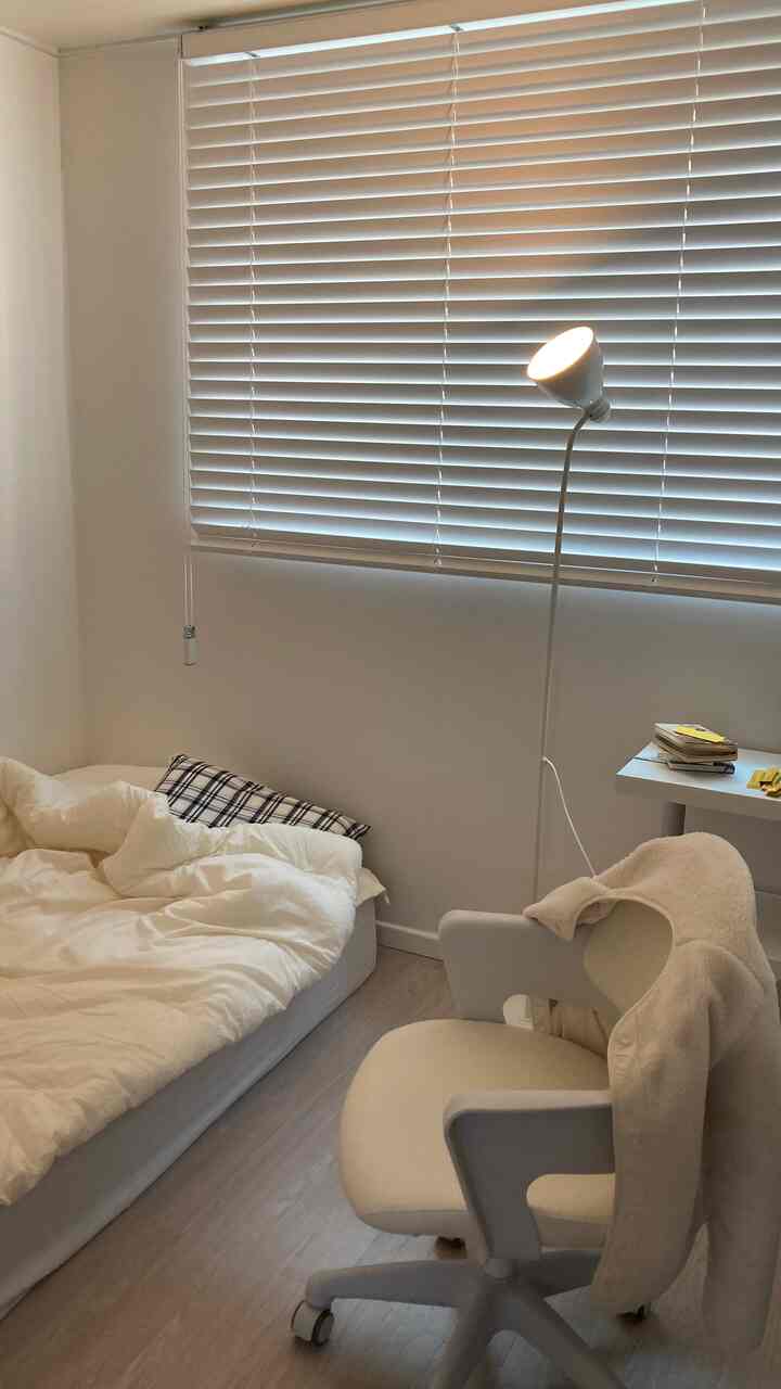 A white and wood-tone compact single household bedroom featuring a desk and floor lamp, creating a cozy home office and sleeping area