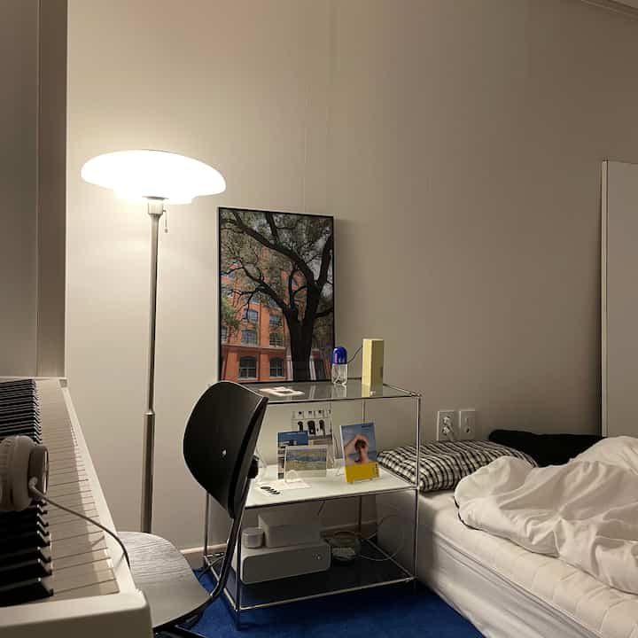 白い壁とベッドがあるモダンなワンルーム、フロアランプと机が配置された落ち着いた空間