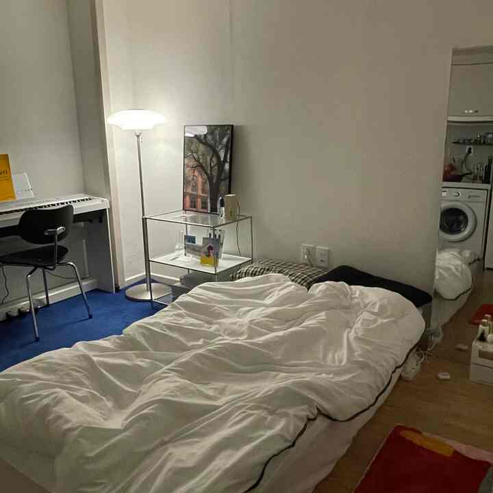 A white and blue toned studio apartment bedroom featuring a bed, piano desk, and floor lamp with cozy atmosphere