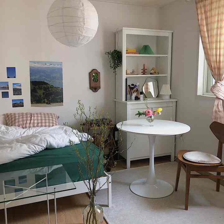 A natural and cozy bedroom-living room featuring white and wood tones with mattress, round dining table, and bookshelf arranged harmoniously.