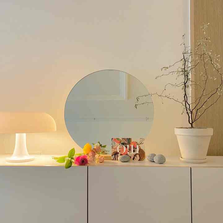 A clean and modern living room setup featuring an ivory-white cabinet with a round mirror, table lamp, and potted plant