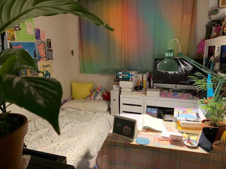 Narrow single bedroom with white walls and rainbow gradient curtain, featuring desk, bed and cozy decor