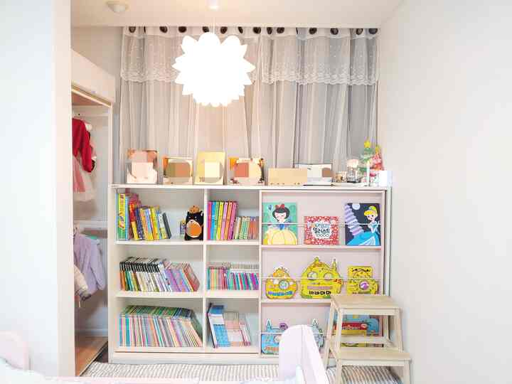 White and beige toned kids' room featuring a central bookshelf, wooden stool, and blackout curtains creating a cozy atmosphere