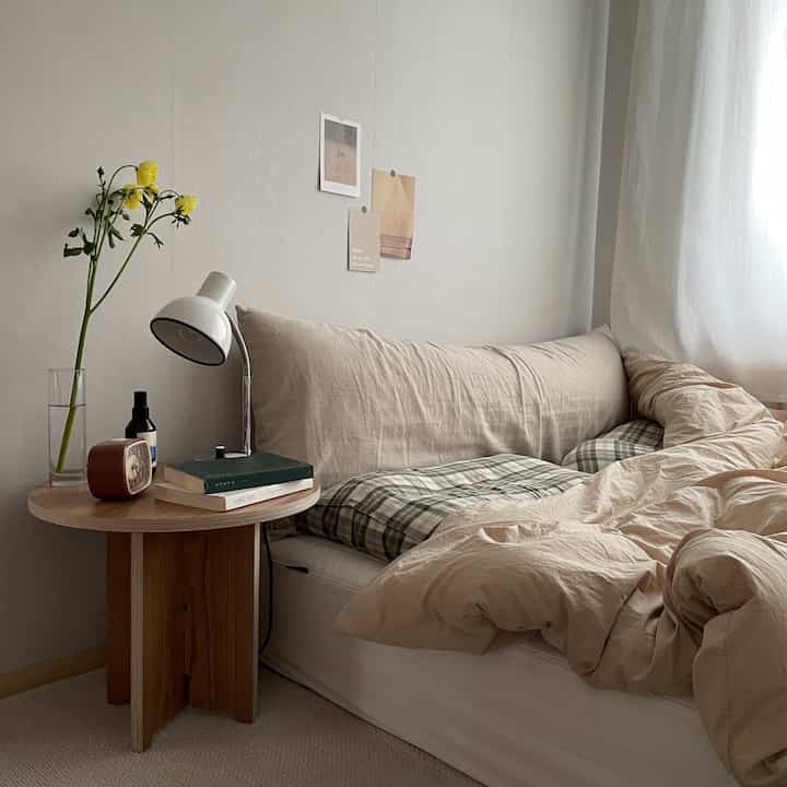 Beige and natural toned bedroom featuring a bed with plaid duvet and round wooden nightstand, creating a cozy atmosphere
