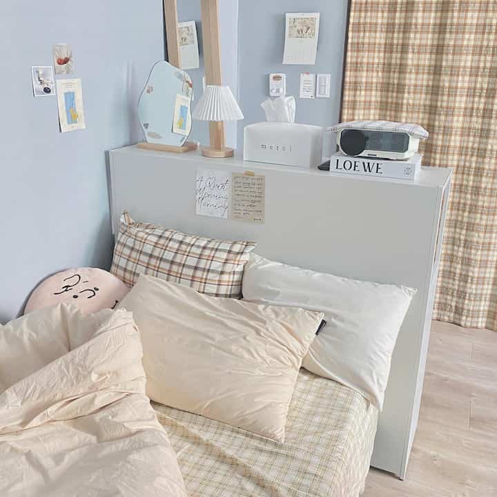 Pastel blue-walled bedroom featuring beige check bedding, a bookshelf serving as bed head, with cozy mood lamp and projector placed on top
