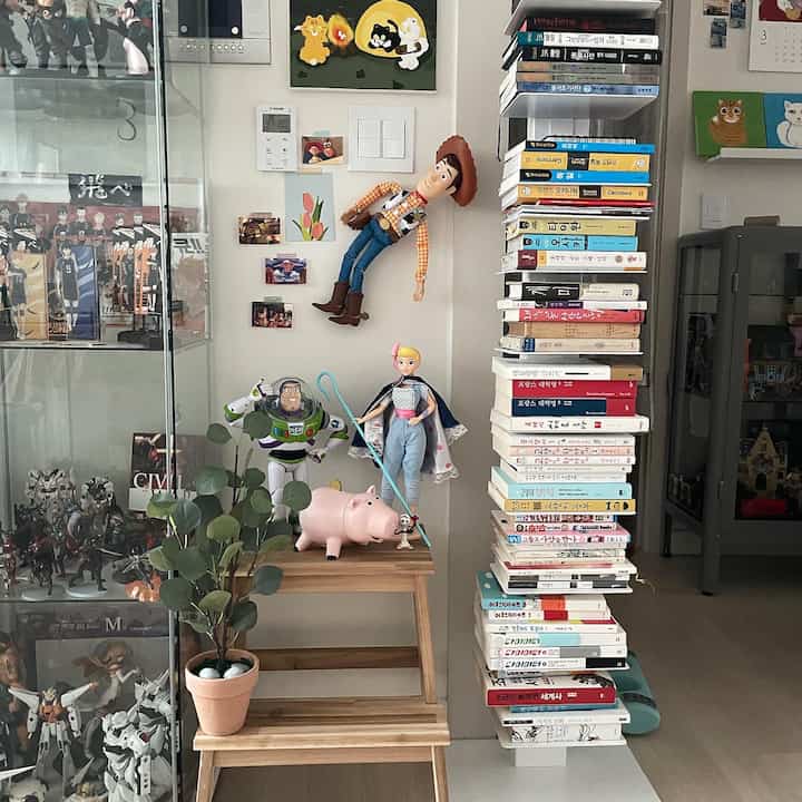 A wood-toned and white wall area featuring a storage stool with dolls, toys, and an artificial plant, creating a vintage atmosphere